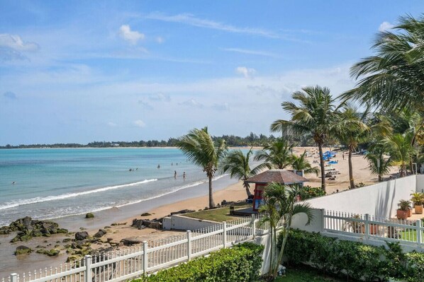 A beach view with a white fence, palm trees, and a small structure near the water. - Beachfront Sanctuary Newly Renovated to Perfection with Full Power Generator (Carolina)