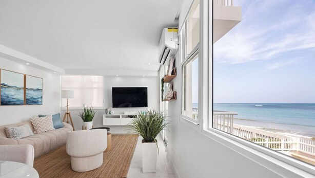 A modern living room with a large window overlooking a beach, a flat-screen TV, and a cozy seating area.