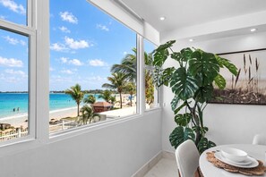 Een kamer met een groot raam met uitzicht op een strand, een eethoek met een tafel en stoelen, en een grote plant.