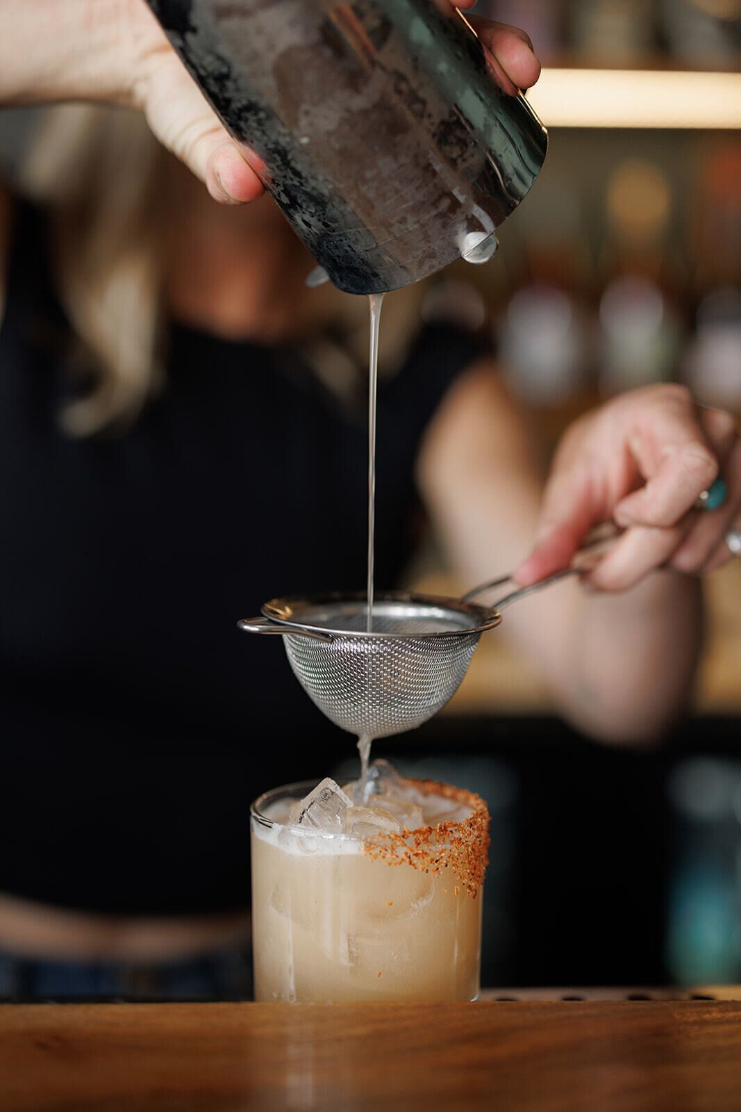 A person pouring a clear liquid into a glass with ice and a rim of orange spice.