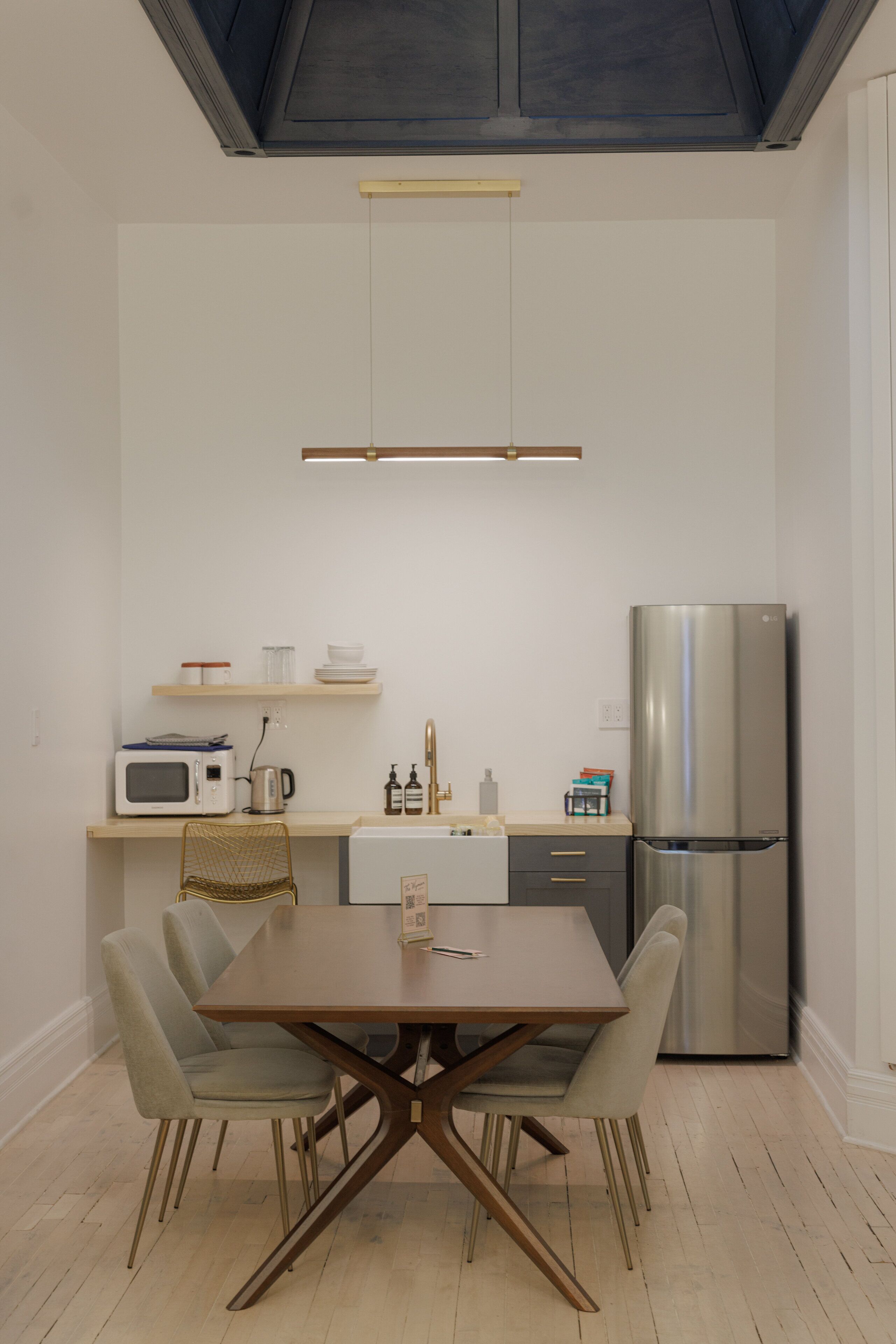 Una cocina moderna con una mesa de comedor, sillas, microondas, hervidor, refrigerador y un estante con vajilla.