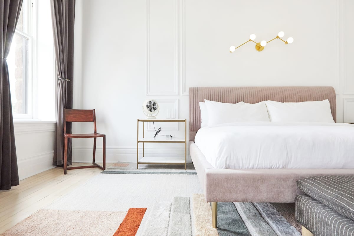 A bedroom with a bed, bedside table, chair, and a small table with a fan.