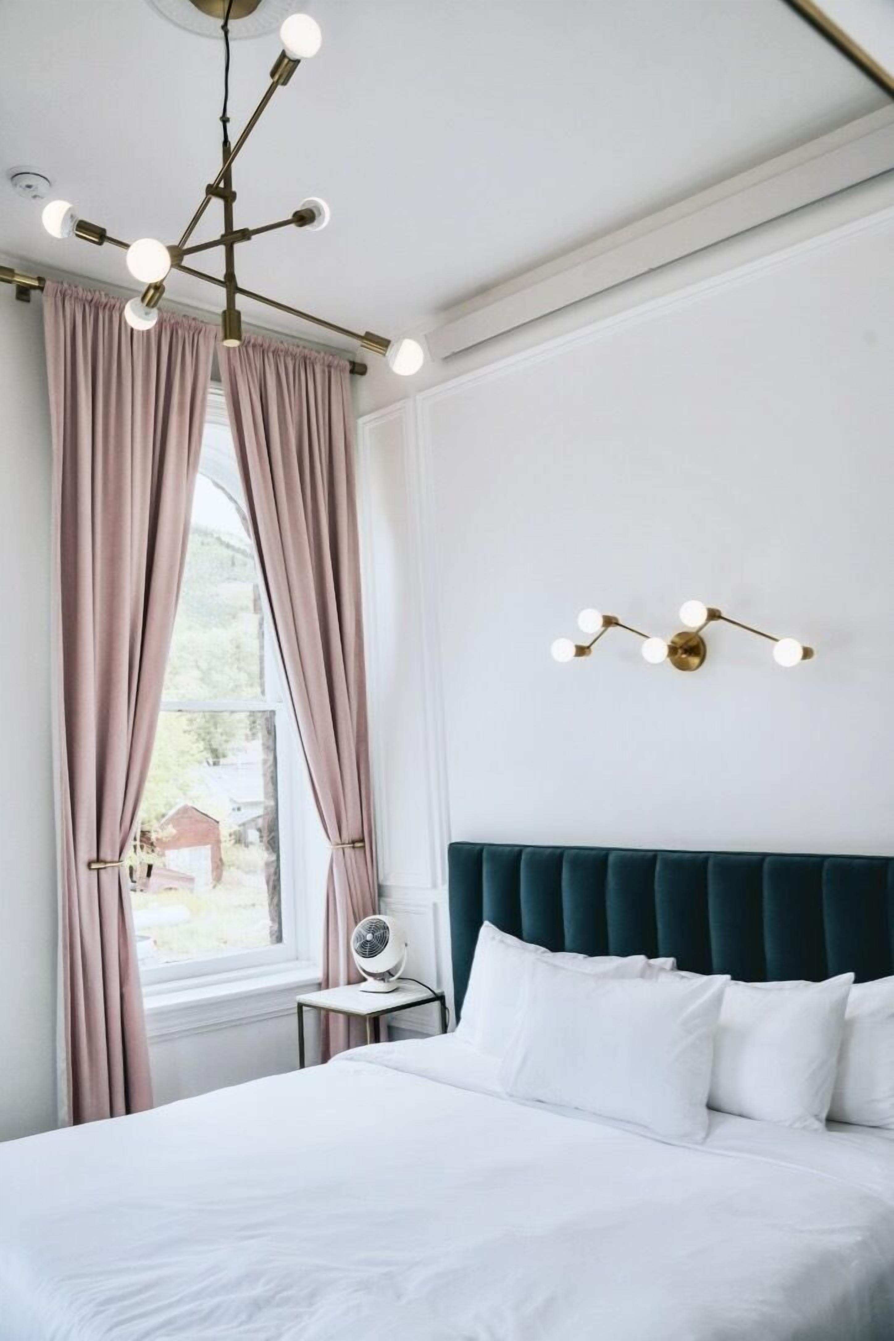 Ein Schlafzimmer mit einem Bett, einem Nachttisch, einem Ventilator, einem Fenster mit Vorhängen und einer Wandleuchte.