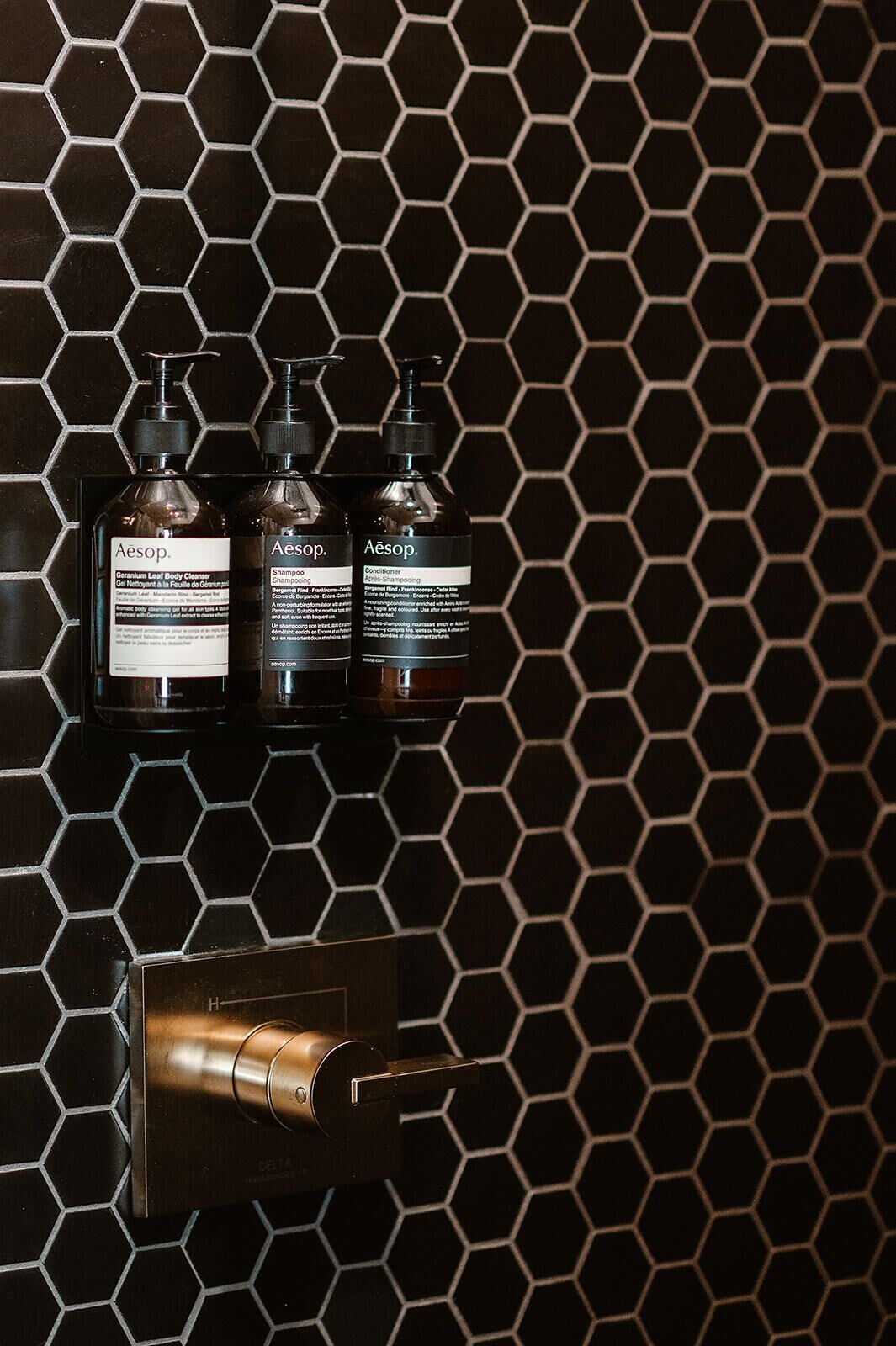 Three bottles of Aesop products on a black hexagonal tile wall.