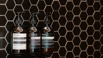 Three bottles of Aesop products on a black hexagonal tile wall.