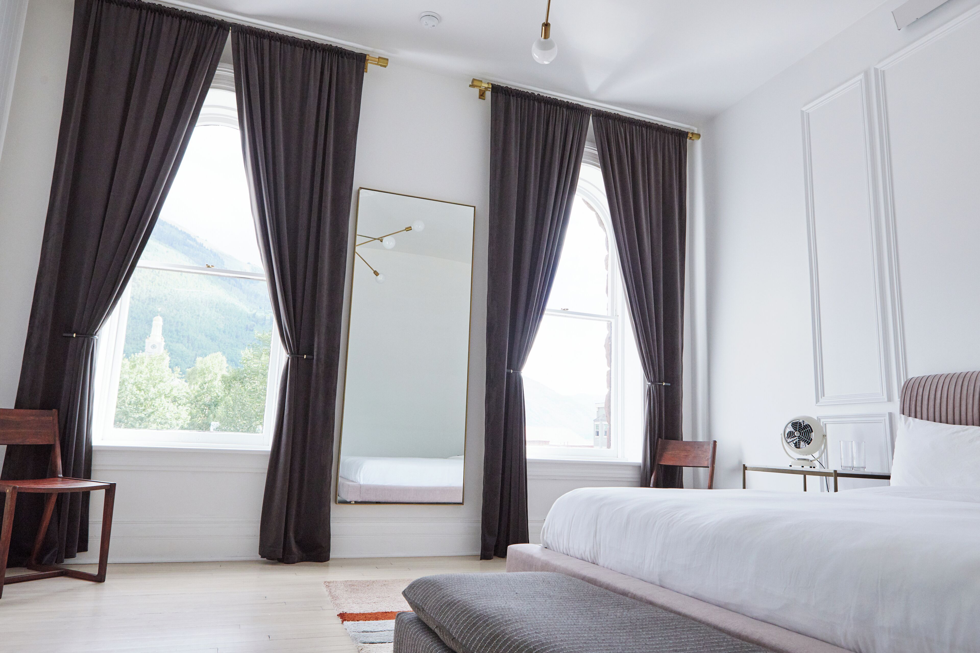 Une chambre avec un grand lit, deux fenêtres avec des rideaux, un miroir, une table de chevet avec une lampe, et une vue sur la verdure à l’extérieur.