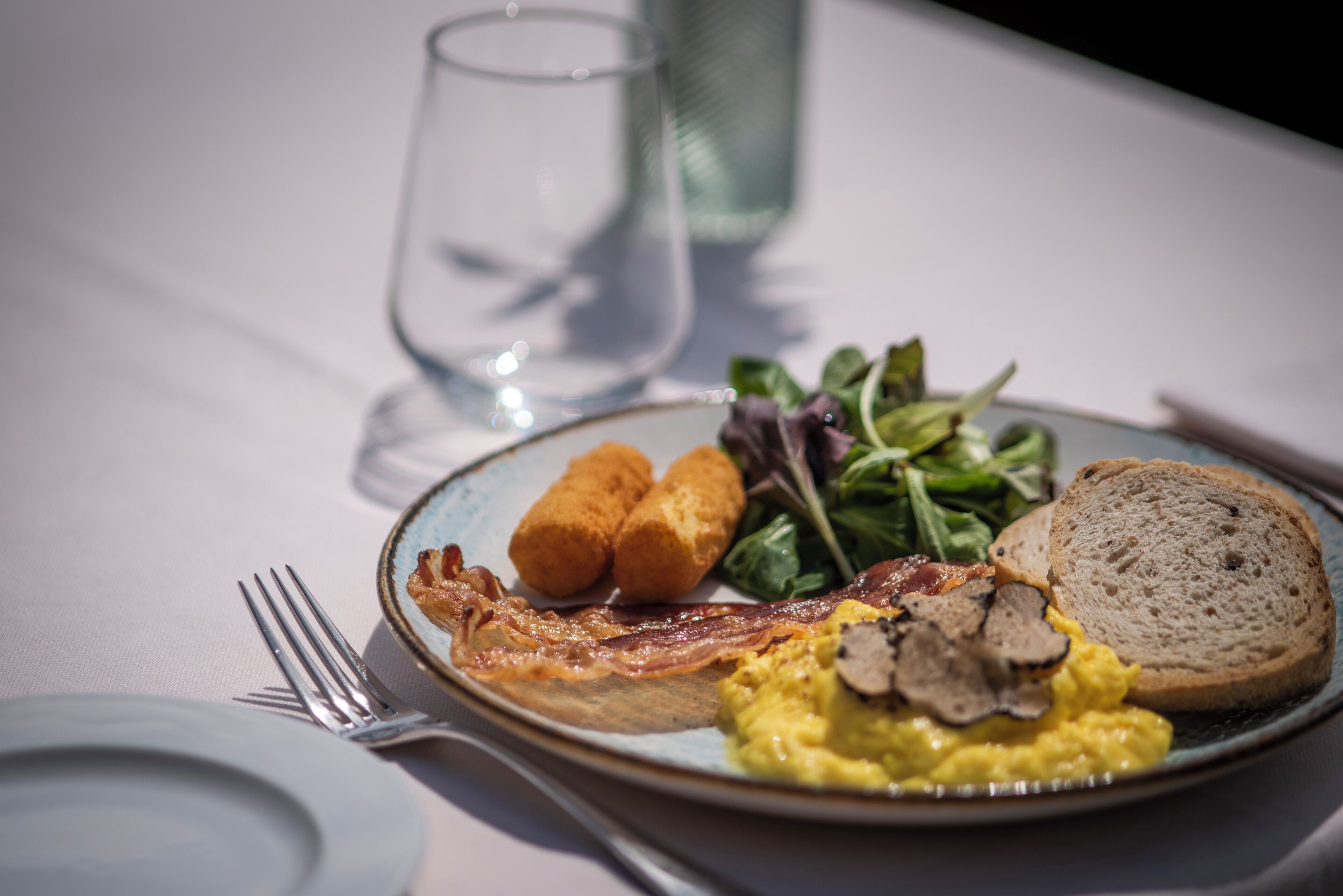 Un plato de desayuno con huevos, tocino, tostadas, vegetales verdes y croquetas.
