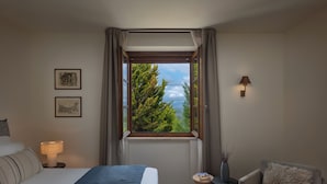 Quarto de hotel com cama, cadeira, luminária, mesa de cabeceira e janela com cortinas.