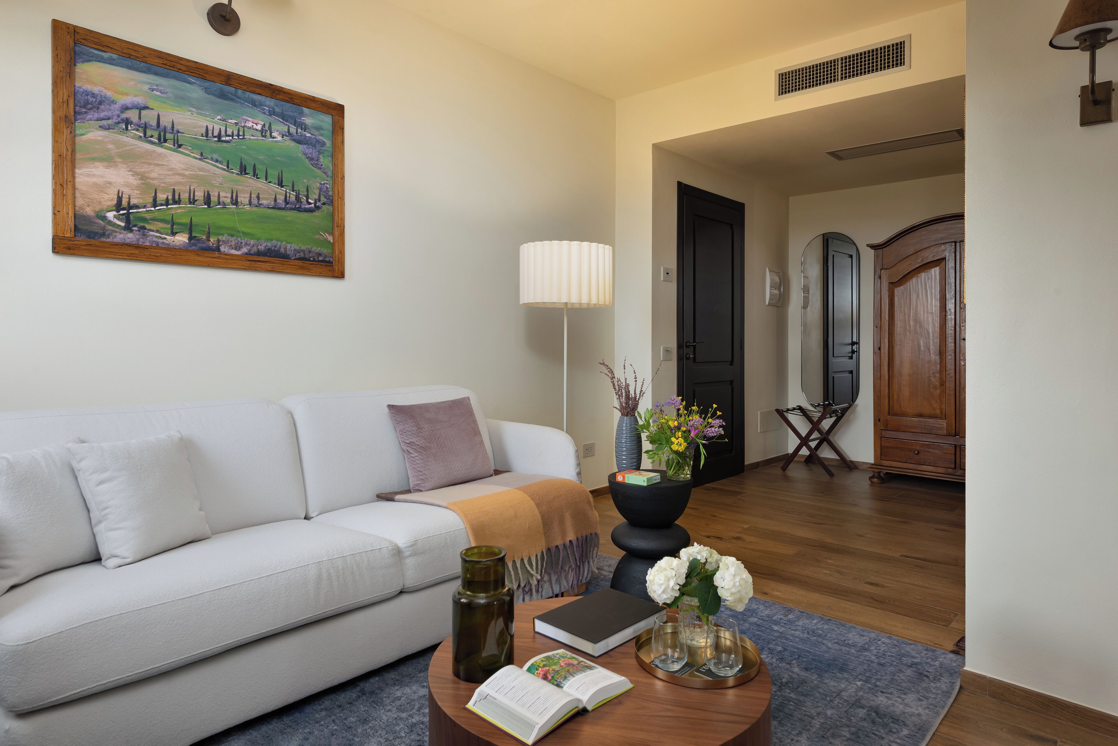 Ein Wohnzimmer mit einem weißen Sofa, einem Gemälde, einer Lampe, einem Couchtisch mit Büchern und Blumen sowie einem Holzkabinett.