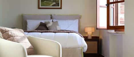 Un dormitorio con una cama, una silla, una mesita de noche con lámpara, una ventana y un cuadro en la pared.