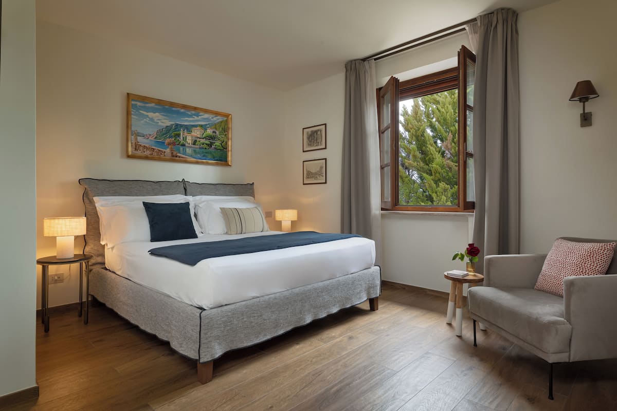Een keurig opgemaakt slaapkamer met een groot bed, nachtlampjes, een klein tafeltje met een vaas en een raam met gordijnen.