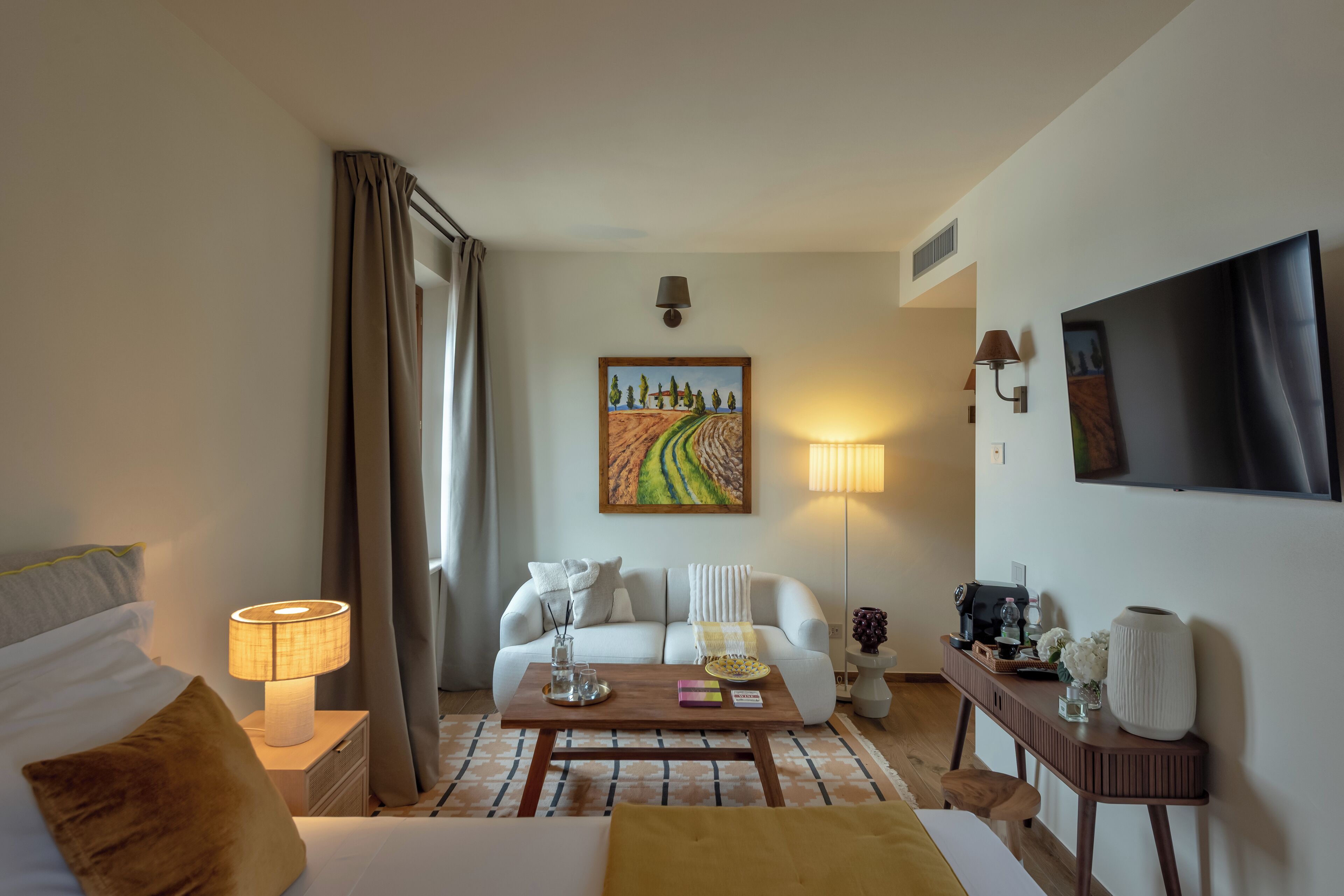 A hotel room with a bed, a sofa, a coffee table, a TV, and a painting on the wall.
