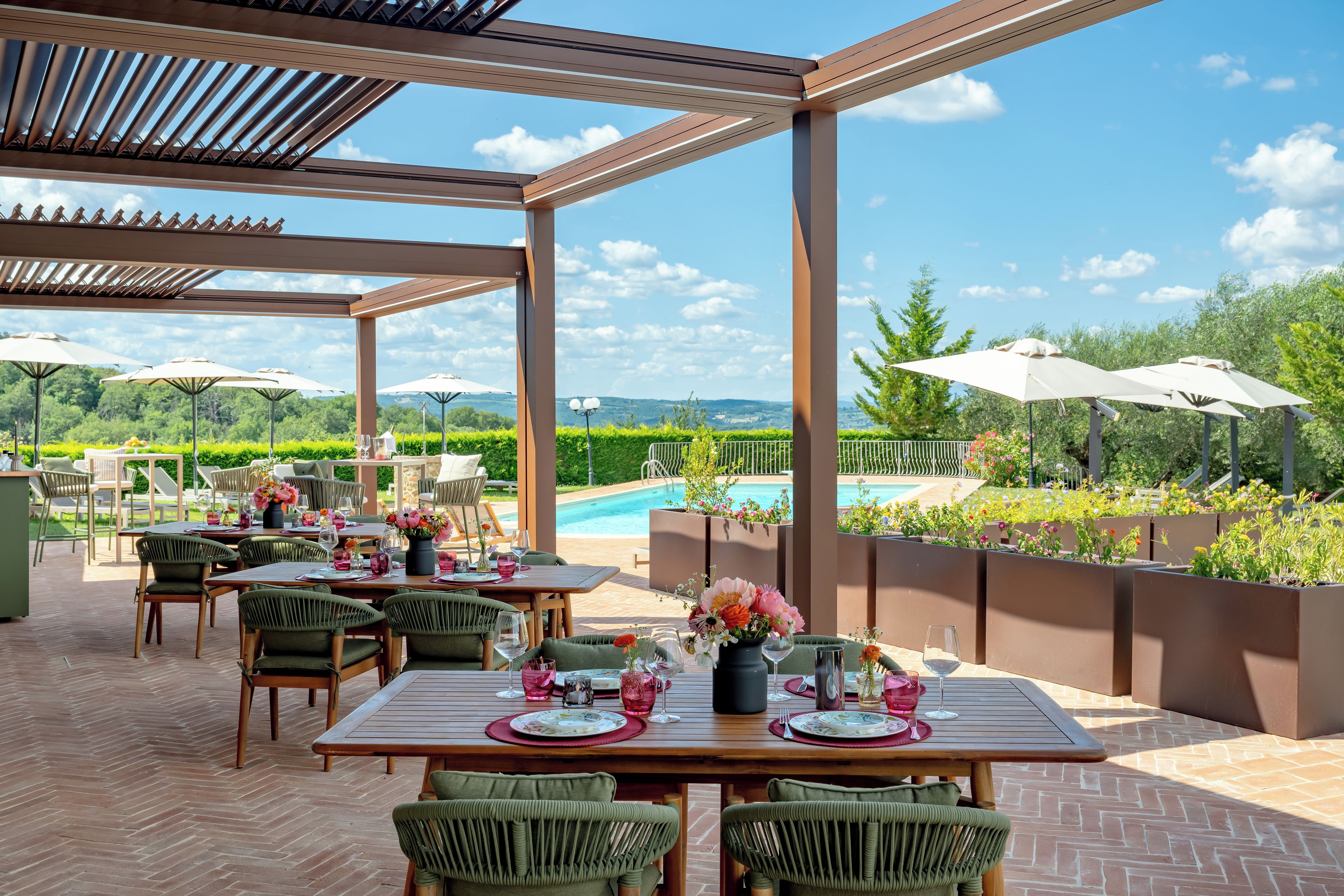 An outdoor dining area with tables and chairs, a pergola, and a view of a swimming pool and greenery.