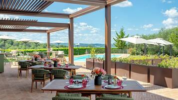 An outdoor dining area with tables and chairs, a pergola, and a view of a swimming pool and greenery.