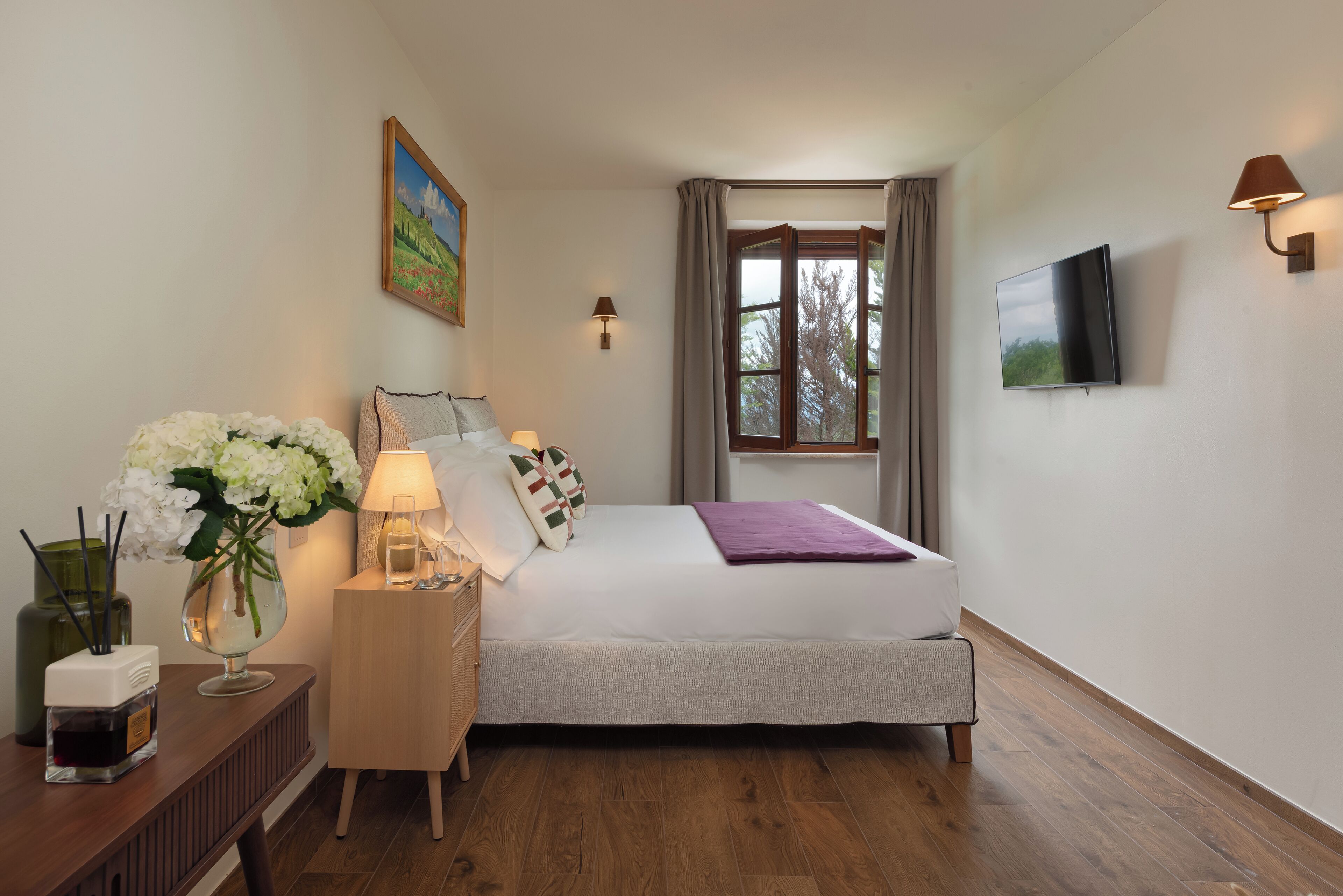 Quarto com cama, mesa de cabeceira, luminária, flores e televisão.