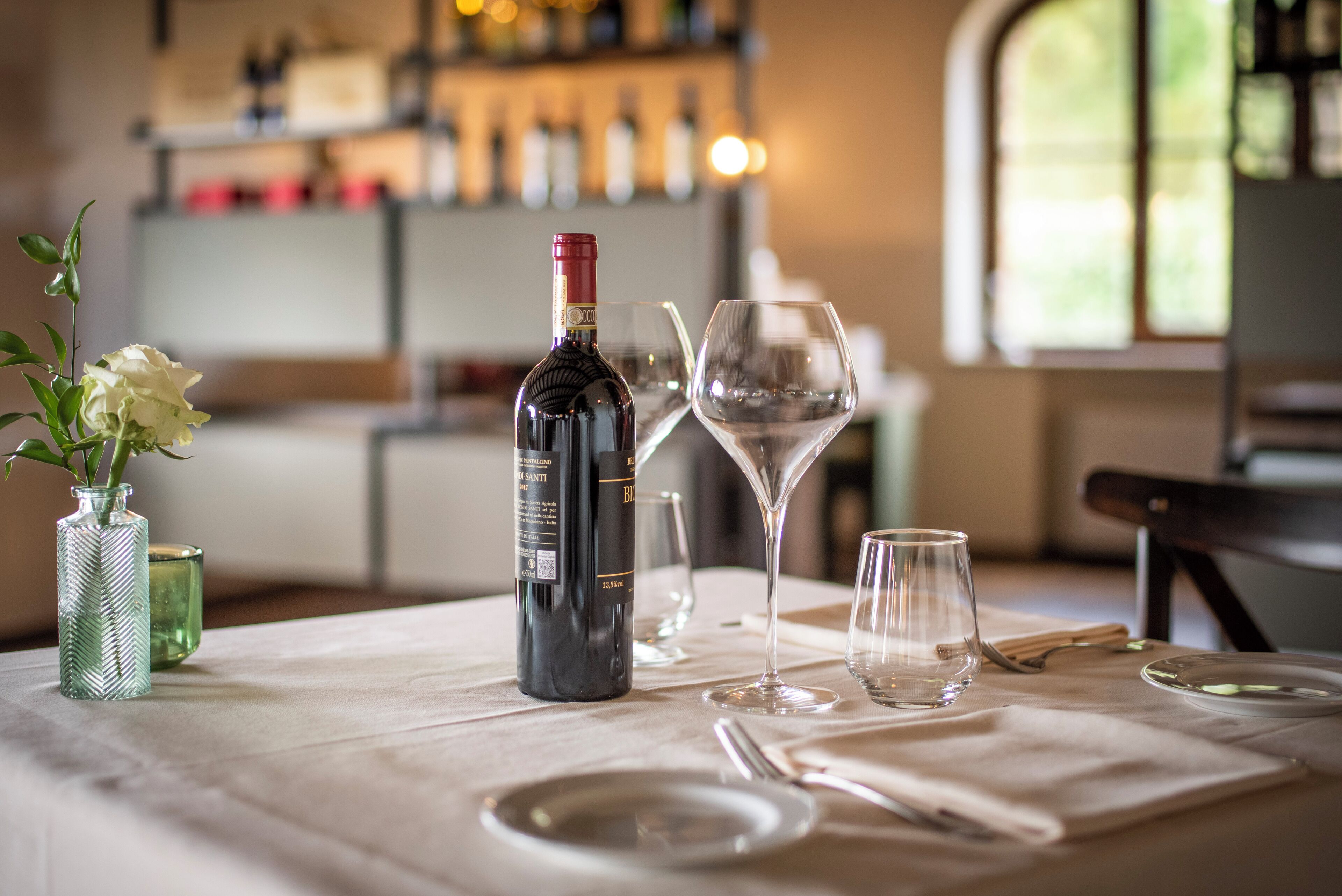 Una mesa dispuesta con una botella de vino, dos copas de vino y un florero con flores.
