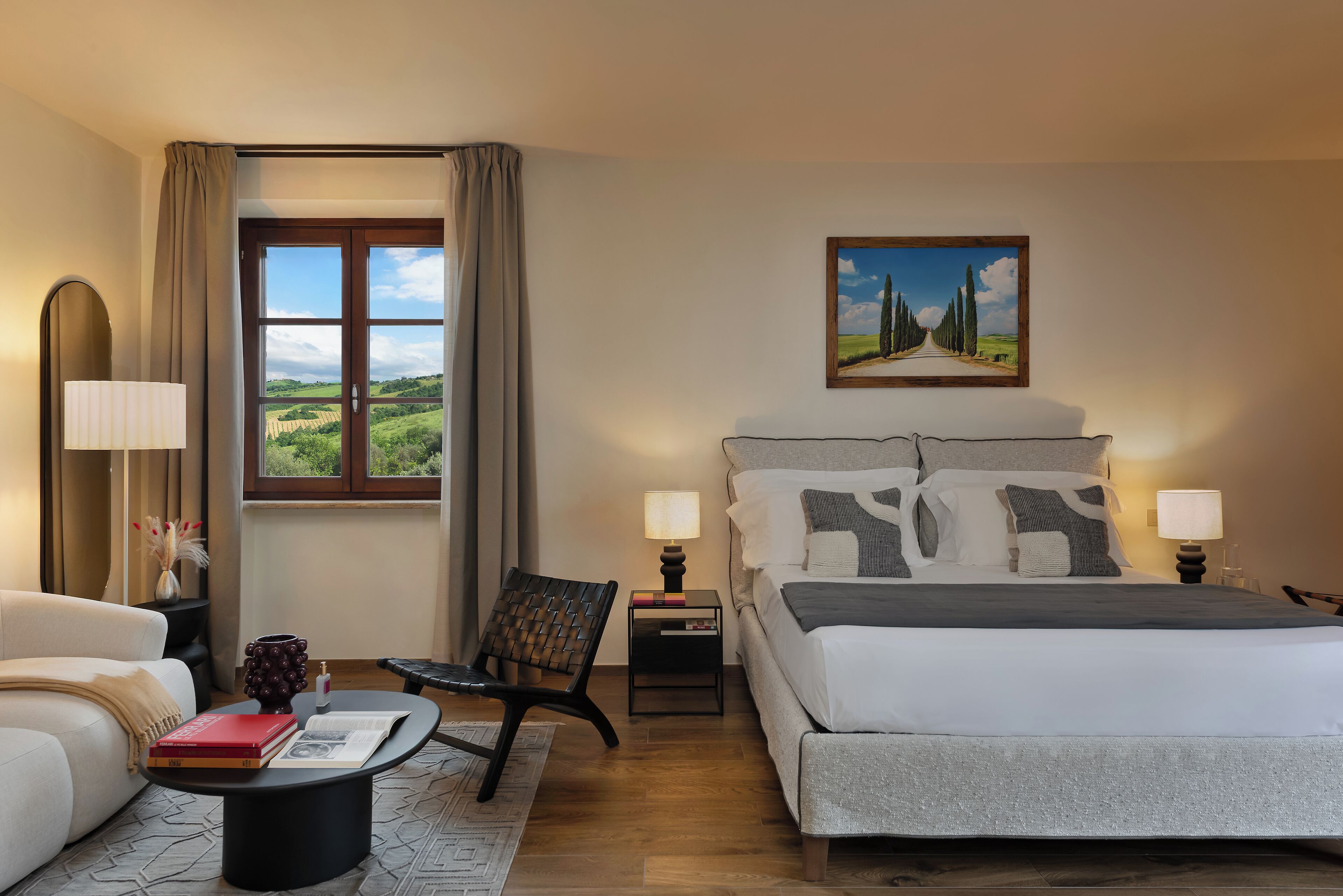 Una habitación de hotel con una cama grande, una silla, una mesita pequeña, una mesita de noche, una lámpara, una ventana con vistas a zonas verdes y un cuadro en la pared.