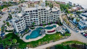 Un hotel de varios pisos con área de piscina, rodeado de vegetación y una playa.