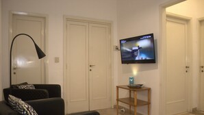 Ein Wohnzimmer mit einem schwarzen Sofa, einem kleinen Holztisch, einer schwarzen Stehlampe und einem flachbildfernseher, der an der Wand montiert ist.
