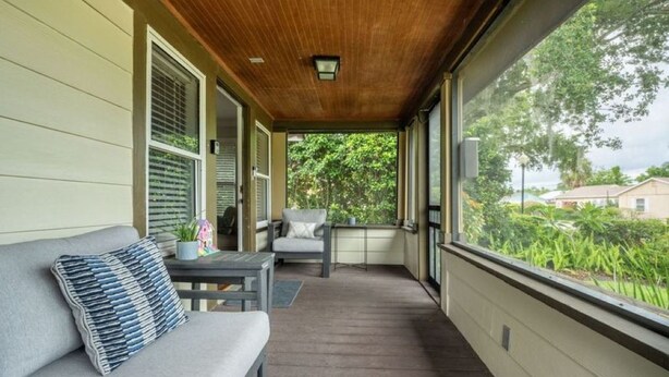 Une véranda couverte avec un plafond en bois, un canapé gris agrémenté d’un coussin à rayures, une petite table et une chaise.