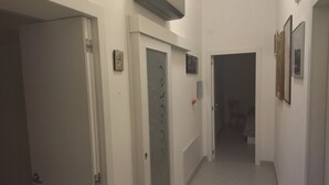 Een smalle gang met witte muren en deuren, die leidt naar een kamer met een bed en een stoel.