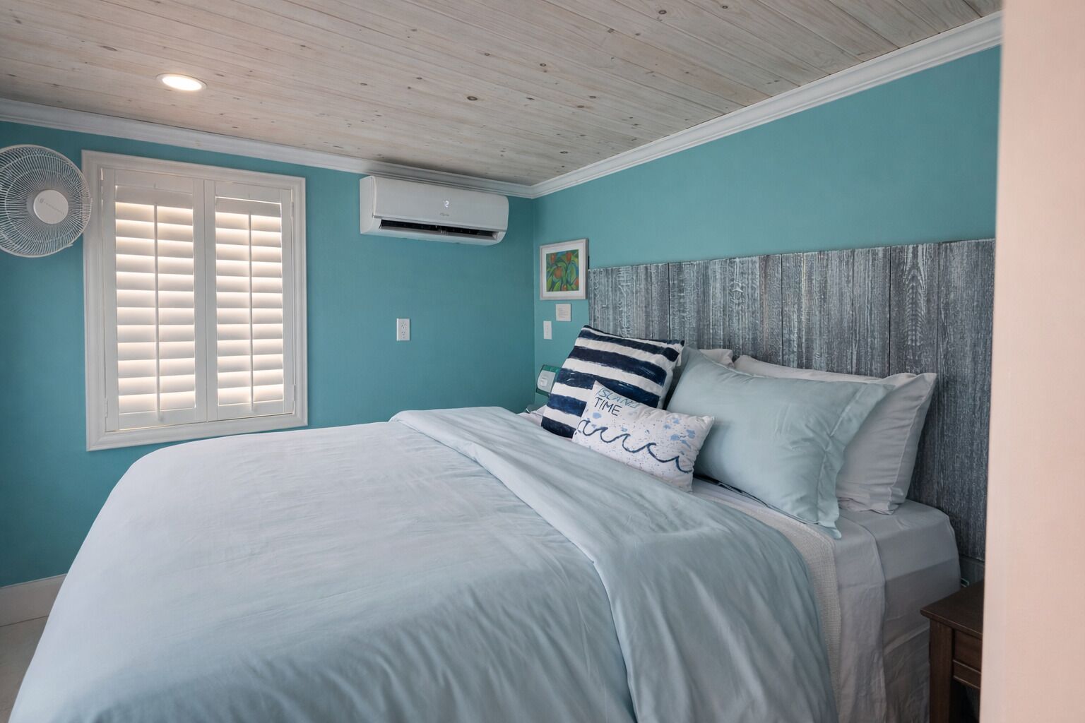 Une chambre à coucher comprenant un lit, un climatiseur fixé au mur, une fenêtre avec des stores et un ventilateur.