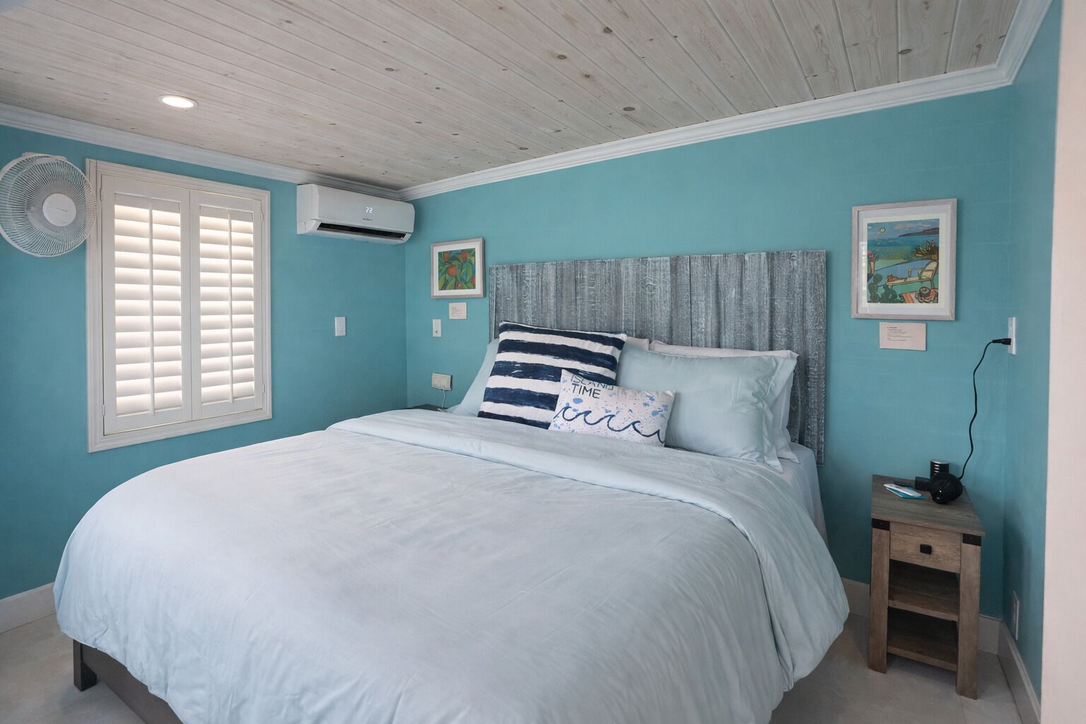 Une chambre à coucher comprenant un lit, un ventilateur sur pied, un appareil de climatisation, une table de chevet et deux tableaux encadrés accrochés au mur.