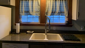 A kitchen with a double sink, a window with plaid curtains, and a countertop with a paper towel holder and a dish rack. - Cozy and relaxing (Linton)