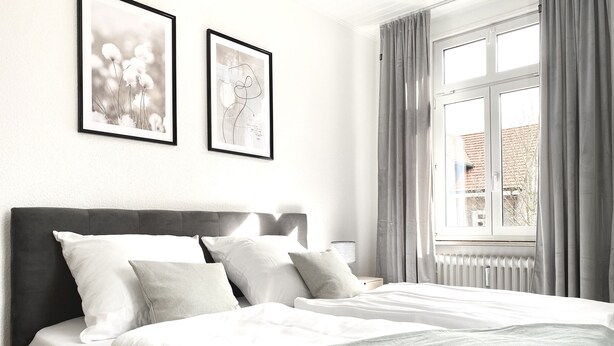 Un lit bien fait, avec du linge de lit blanc et un plaid gris, une tête de lit rembourrée de couleur foncée et deux tableaux encadrés accrochés au mur.