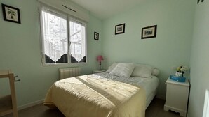 A bedroom with a bed, a bedside table with a lamp, a radiator, and a window with curtains. - South-facing chalet in Erquy | 8 guests | Ref 759 (ERQUY)