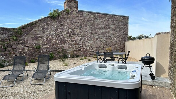 Et boblebad i en baghave med stenvægge, et bord med stole og en grill.