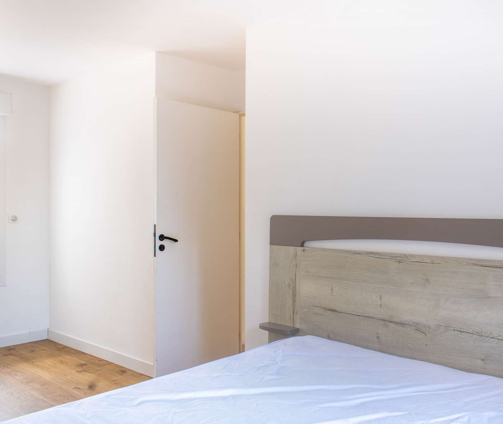 A bedroom with a wooden headboard, a white mattress, and a door leading to another room.