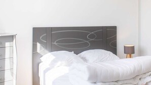 En pænt redt seng med hvidt sengetøj, et moderne abstrakt kunstværk på væggen over sengen, og et natbord med en lampe.