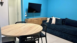 Ein Zimmer mit einem runden Holztisch, schwarzen Stühlen, einem dunkelblauen Sofa mit Kissen, einem wandmontierten Flachbildfernseher und einem Holzschrank.
