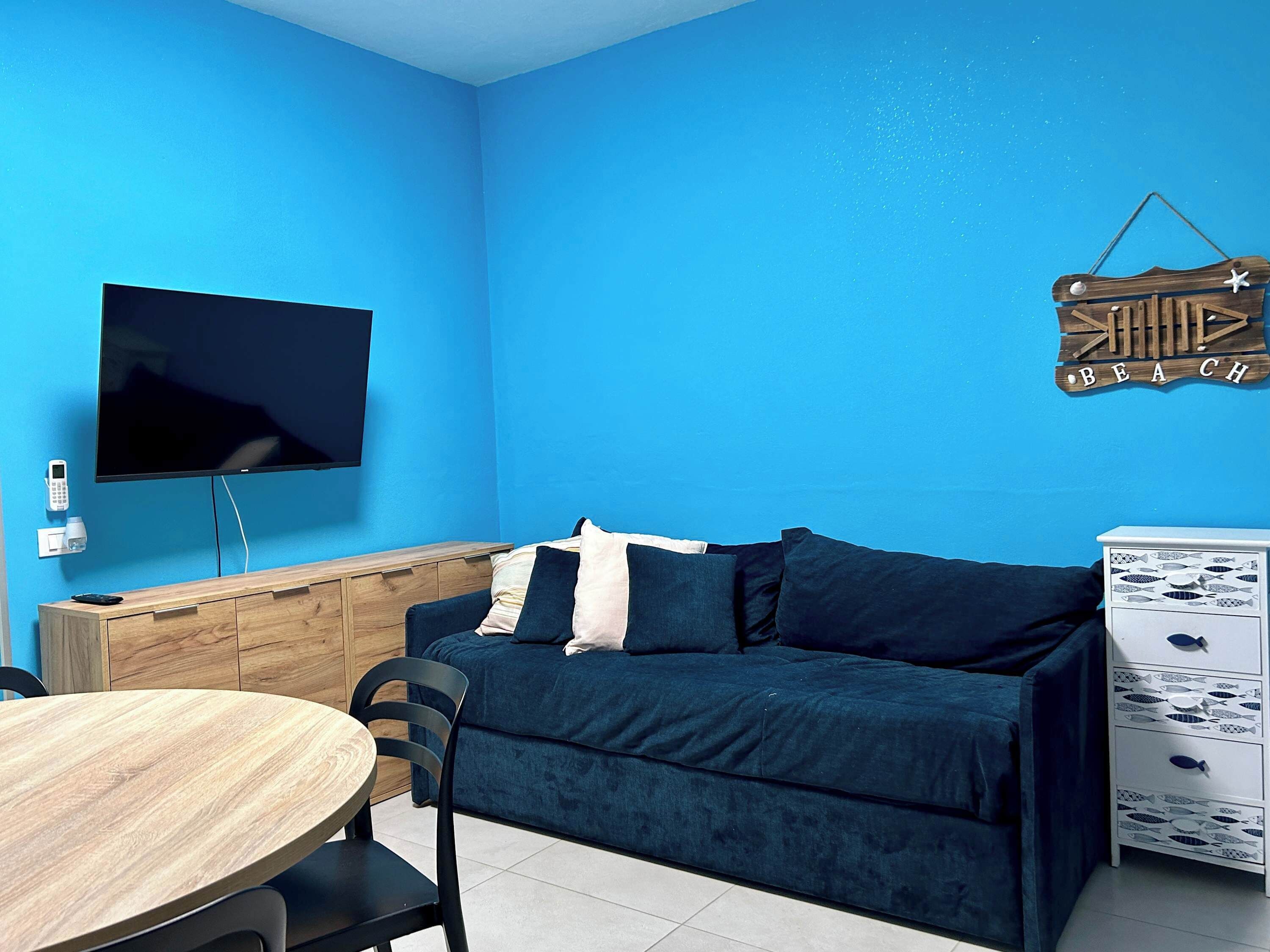 A room with a blue wall, a flat-screen TV mounted on the wall, a wooden cabinet, a dark blue sofa with pillows, a round wooden table with chairs, and a white dresser with patterned drawers.