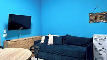 A room with a blue wall, a flat-screen TV mounted on the wall, a wooden cabinet, a dark blue sofa with pillows, a round wooden table with chairs, and a white dresser with patterned drawers.