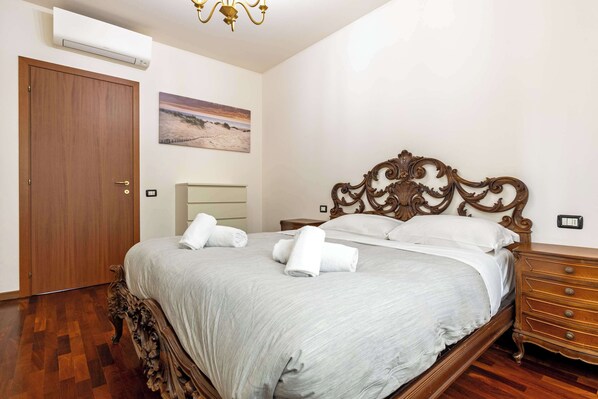 A bedroom with a wooden bed, a nightstand, a dresser, a wall-mounted air conditioner, and a painting.