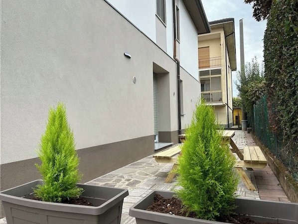 Un estrecho pasillo exterior con dos coníferas en macetas, un camino de piedra y un banco de madera.