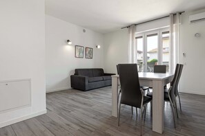 A modern living room with a grey sofa, a dining table with four chairs, and a large window with curtains.