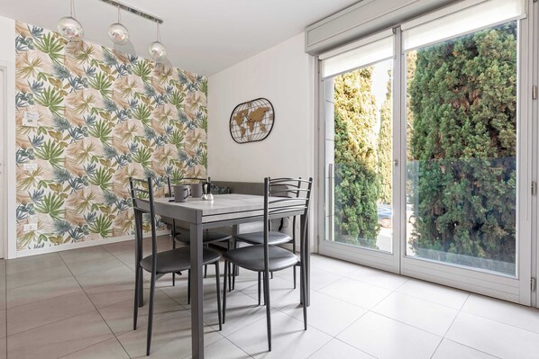 A dining area with a table and chairs, a world map wall decoration, and a view of a green hedge through a glass door.