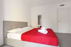A neatly made bed with a red bedspread and white pillows, a grey headboard, and a white door in the background. - Vivaldi Home - Immobili e Soluzioni Rent (Bergamo)