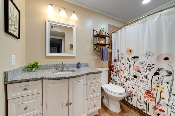 A bathroom with a granite countertop sink, a white toilet, a shower curtain with floral patterns, a mirror above the sink, and a shelf with a potted plant.