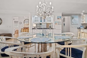 A dining area with a glass table surrounded by white chairs with blue cushions, a chandelier, and a kitchen in the background.