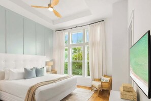 A bedroom with a large bed, a ceiling fan, a window with curtains, a television, and a small table with a chair.