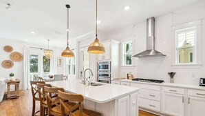 A modern kitchen with white cabinetry, a central island with bar stools, pendant lighting, and a stainless steel range hood.