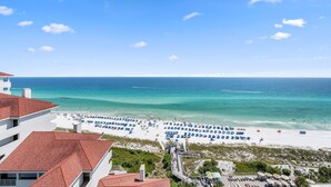 A beach with white sand and clear blue water, a pier, and buildings with red roofs.