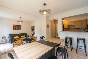A modern living room with a wooden dining table, black chairs, a black sofa with yellow cushions, a bookshelf, and a kitchen area with wooden cabinets and a bar stool.