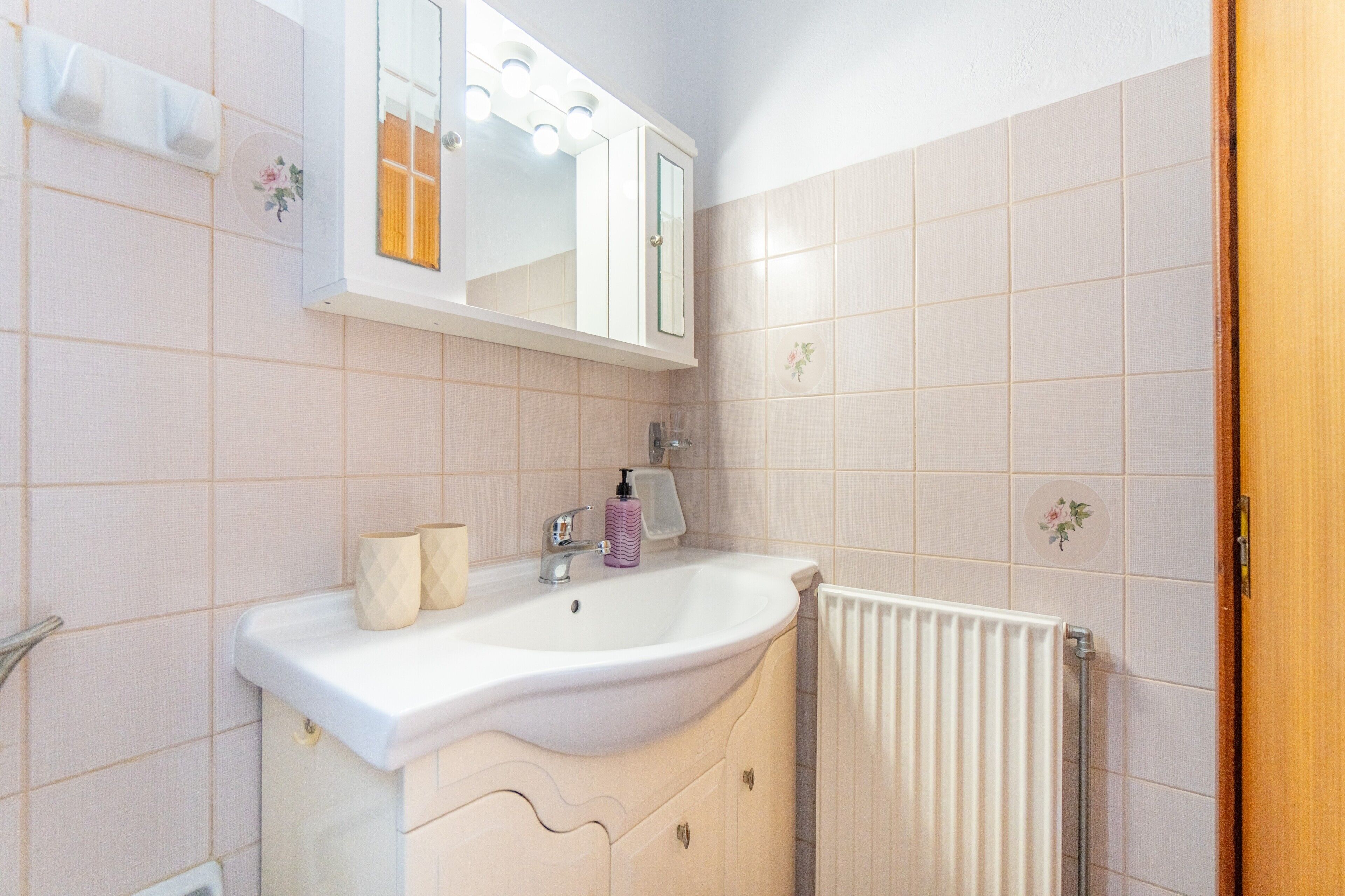 Un baño con lavamanos blanco, espejo y mueble, un radiador y una puerta de madera.