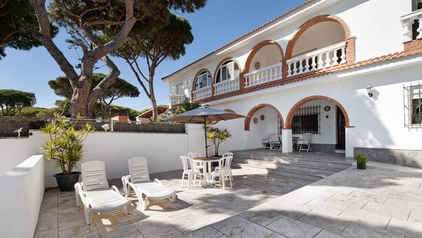 A patio with white chairs and a table, a large tree, and a building with arches and a balcony. - Villa 'El Chozo' with Sea View, Private Pool and Wi-Fi (Chiclana de la Frontera)