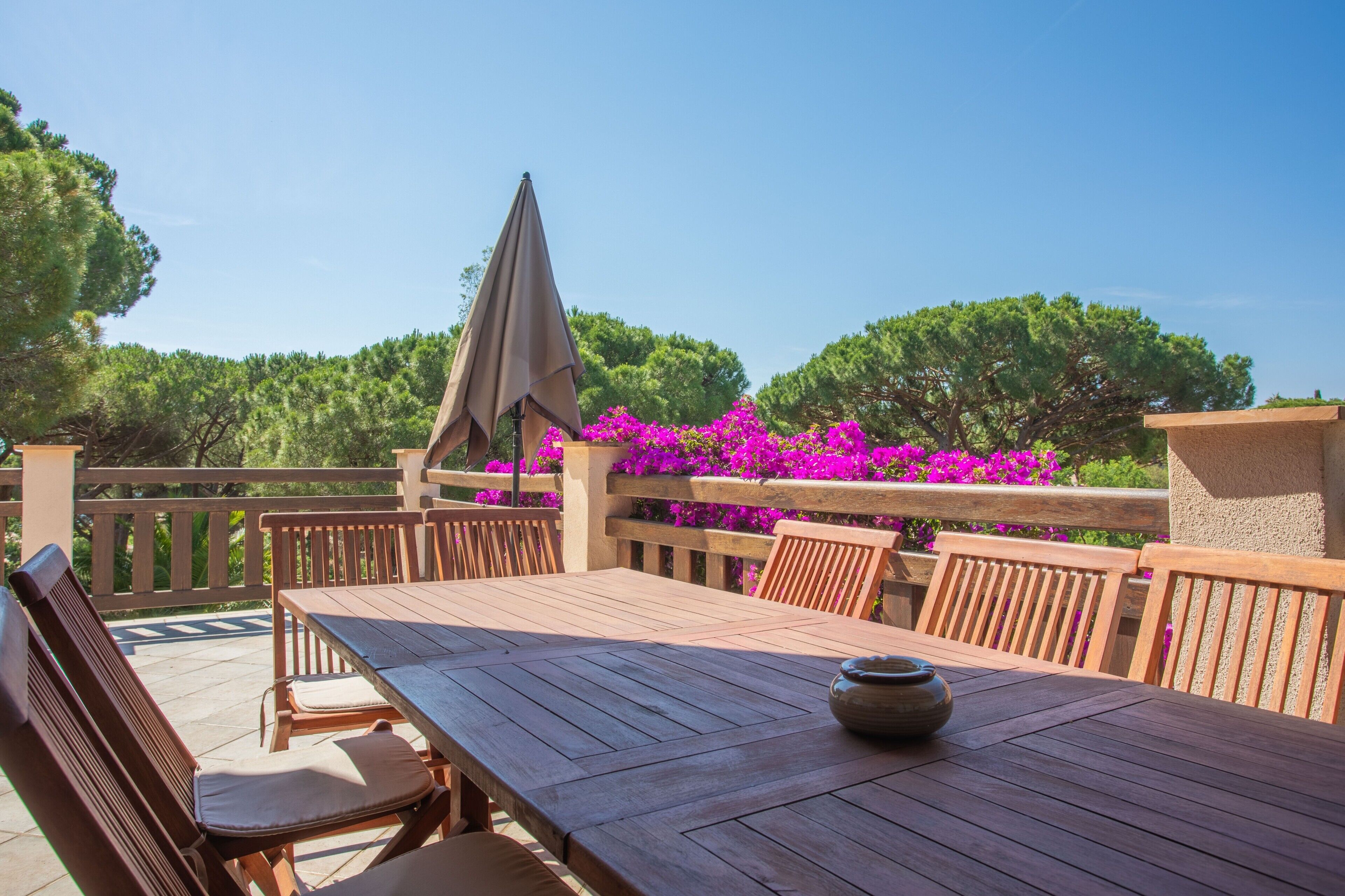 A wooden deck with a table and chairs, surrounded by a wooden railing and vibrant pink flowers.